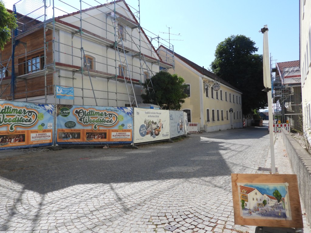 Eine Straße mit einem im Bau befindlichen Gebäude, umgeben von Baugerüsten und Werbebannern für eine Oldtimer-Veranstaltung. Im Vordergrund steht eine Malerei, die eine Szene der Straße darstellt.
