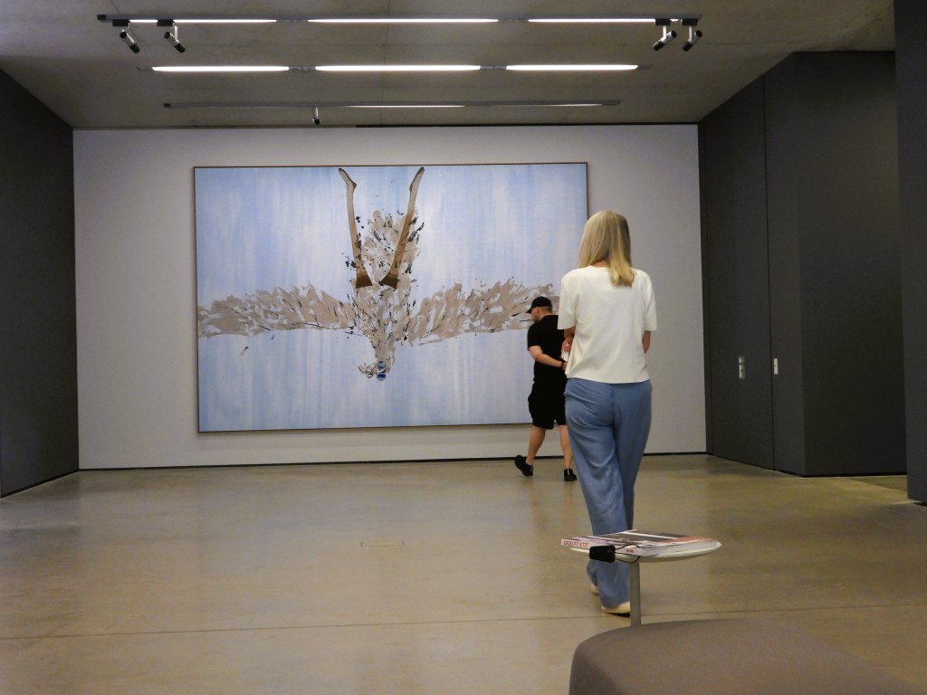 Eine Frau mit langen Haaren in einem weißen Oberteil steht vor einem großen Gemälde in einem modernen Kunstmuseum. Im Hintergrund sind zwei weitere Personen zu sehen.