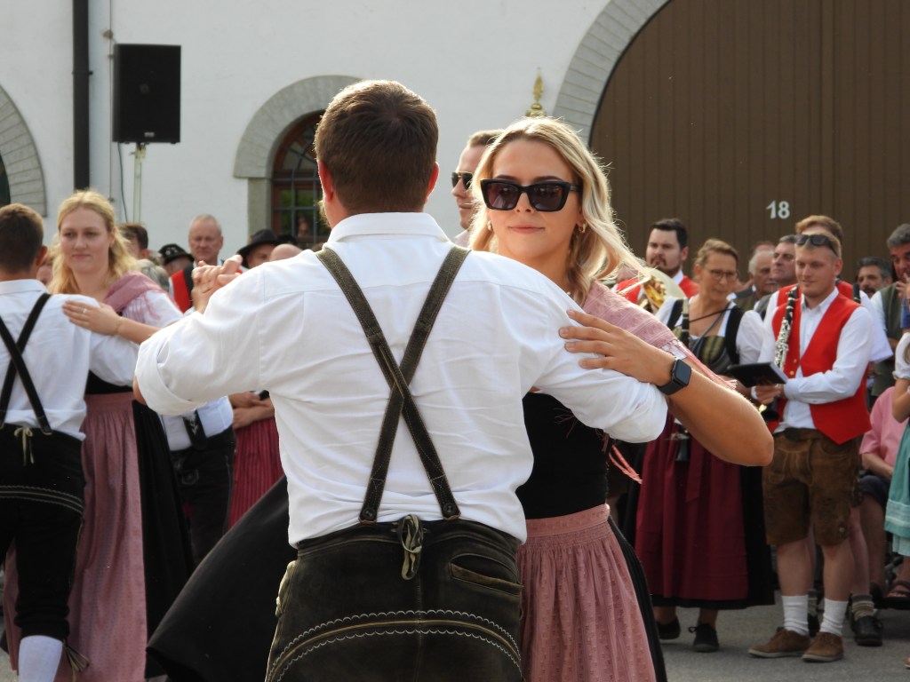 Ein Paar tanzt in traditioneller Kleidung auf einem Fest. Die Frau trägt ein schwarzes Oberteil und einen pinken Rock, während der Mann ein weißes Hemd und Lederhosen an hat. Im Hintergrund sind weitere Tänzer und Zuschauer zu sehen.