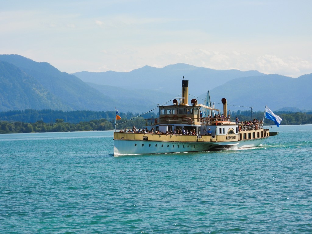 Ein nostalgisches Dampfschiff fährt auf einem klaren See, umgeben von Bergen und einer blauen Himmel.