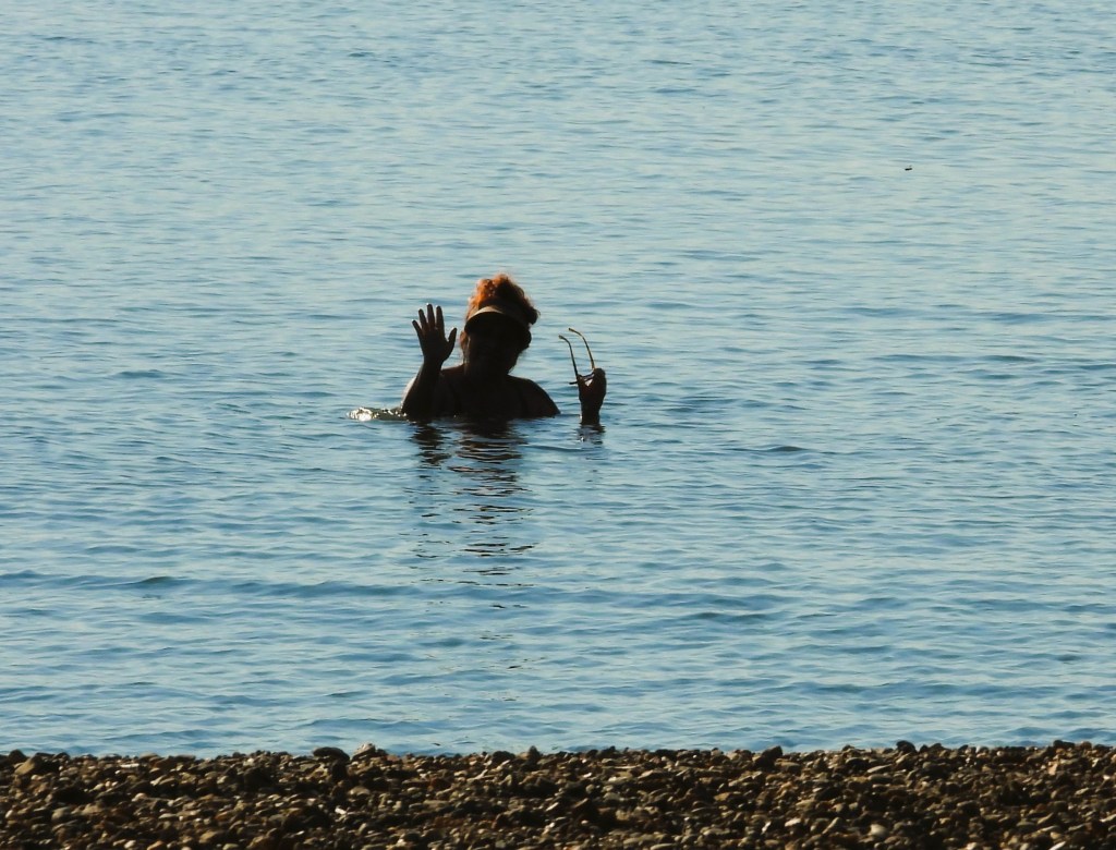 Eine Person steht im Wasser und winkt, während sie von einem Kiesstrand aus betrachtet wird.