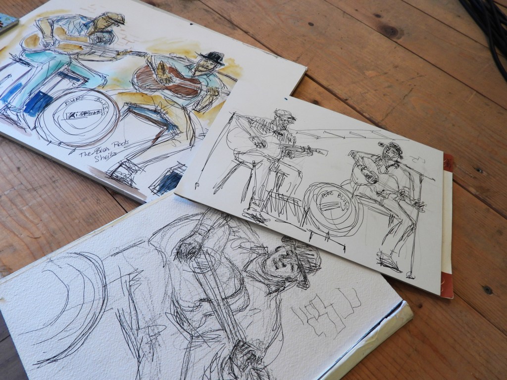 Skizzen von Musikern, die Gitarre spielen, auf verschiedenen Blättern Papier, verteilt auf einem Holztisch.