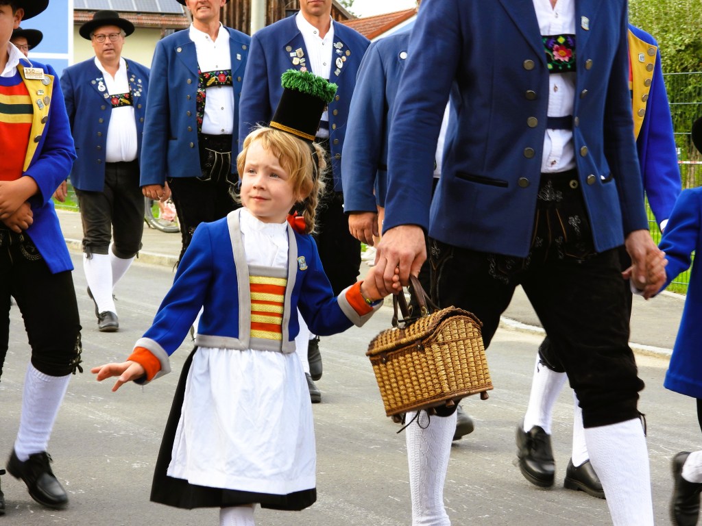 Ein Kind in traditioneller Tracht hält die Hand eines Erwachsenen und läuft an einer Gruppe von Männern in Festtagkleidung vorbei. Die Männer tragen blaue Jacken und Hosen, während das Kind ein weißes Kleid mit einer bunten Jacke trägt. Im Hintergrund sind Häuser und Bäume sichtbar.