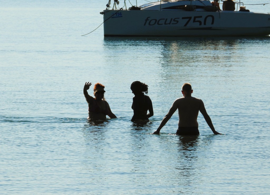 Drei Personen stehen im Wasser, während ein Boot im Hintergrund zu sehen ist.