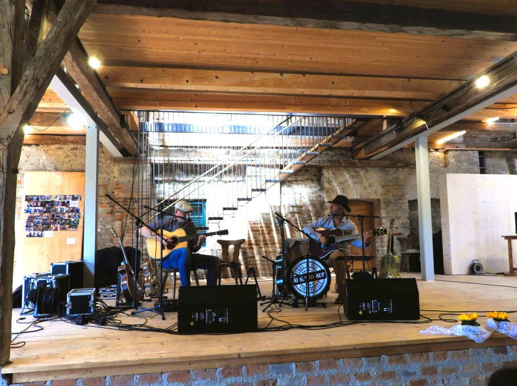 Zwei Musiker spielen auf einer Bühne in einem rustikalen Raum. Der erste Musiker spielt eine akustische Gitarre, während der zweite einen Hut trägt und ebenfalls Gitarre spielt. Im Hintergrund sind Holzstufen und eine Wand mit Fotos zu sehen.