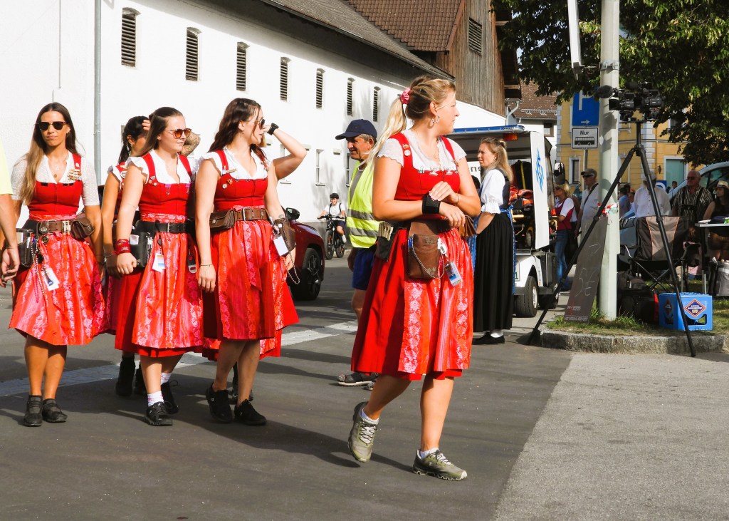 Eine Gruppe von Frauen in roten traditionellen Kleidern, die bei einem Festumzug in einer Stadt gehen. Im Hintergrund sind verschiedene Zuschauer und Fahrzeuge sichtbar.