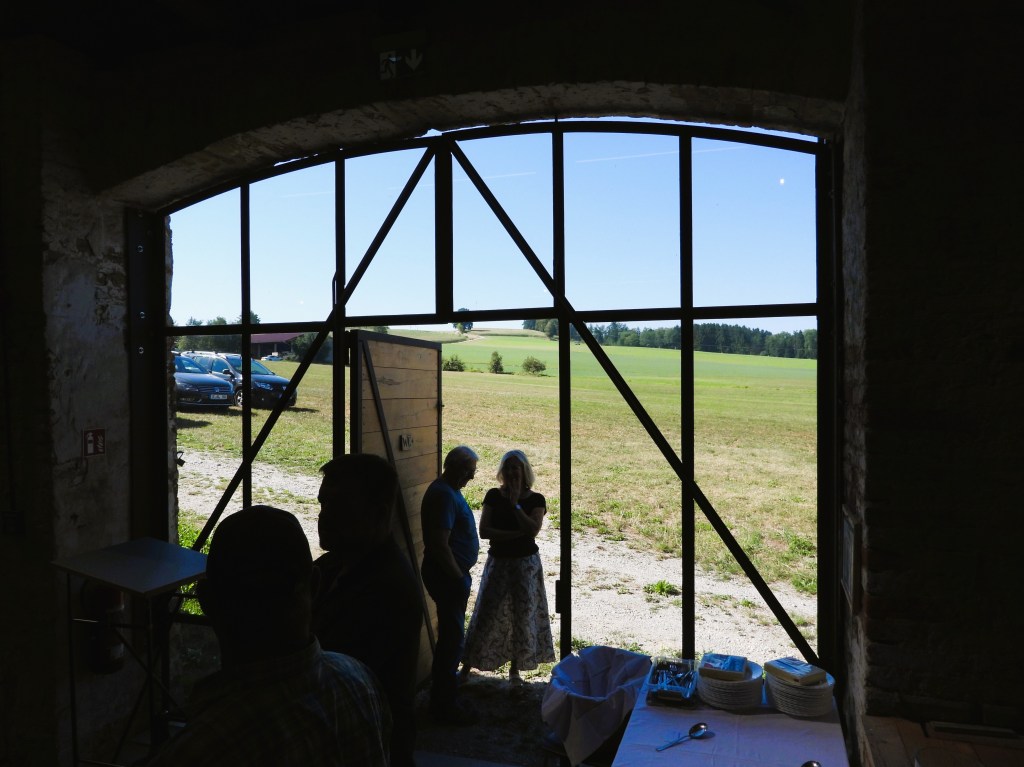Blick aus einem offenen Tor einer Scheune auf eine Wiese und Fahrzeuge im Hintergrund. Im Vordergrund stehen zwei Personen, während Geschirr und Besteck auf einem Tisch vorbereitet werden.