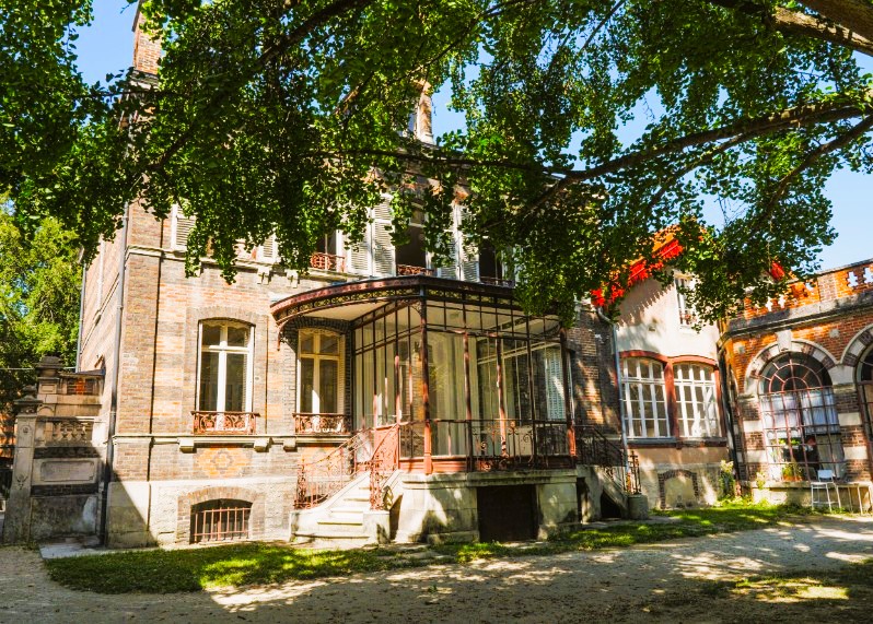 Historisches Gebäude mit roten Ziegelfassade und einem verglasten Erker, umgeben von Bäumen und grünem Gras.