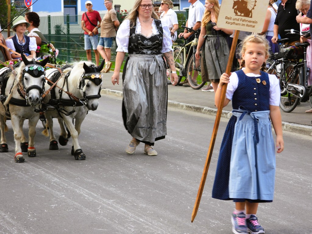 Eine Frau in traditioneller Tracht führt zwei kleine Ponys an, während ein Mädchen mit einer Holztafel im Festumzug geht. Im Hintergrund sind weitere Personen und Fahrräder zu sehen.