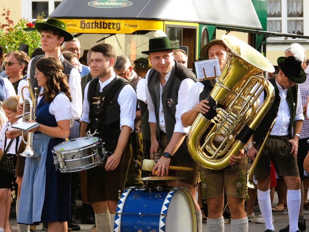 Eine Gruppe von Menschen in traditioneller bayerischer Tracht, die Musikinstrumente halten; eine Frau mit einer Trompete, ein Mann mit einer Tuba und weitere Musiker. Im Hintergrund sind Zuschauer zu sehen.