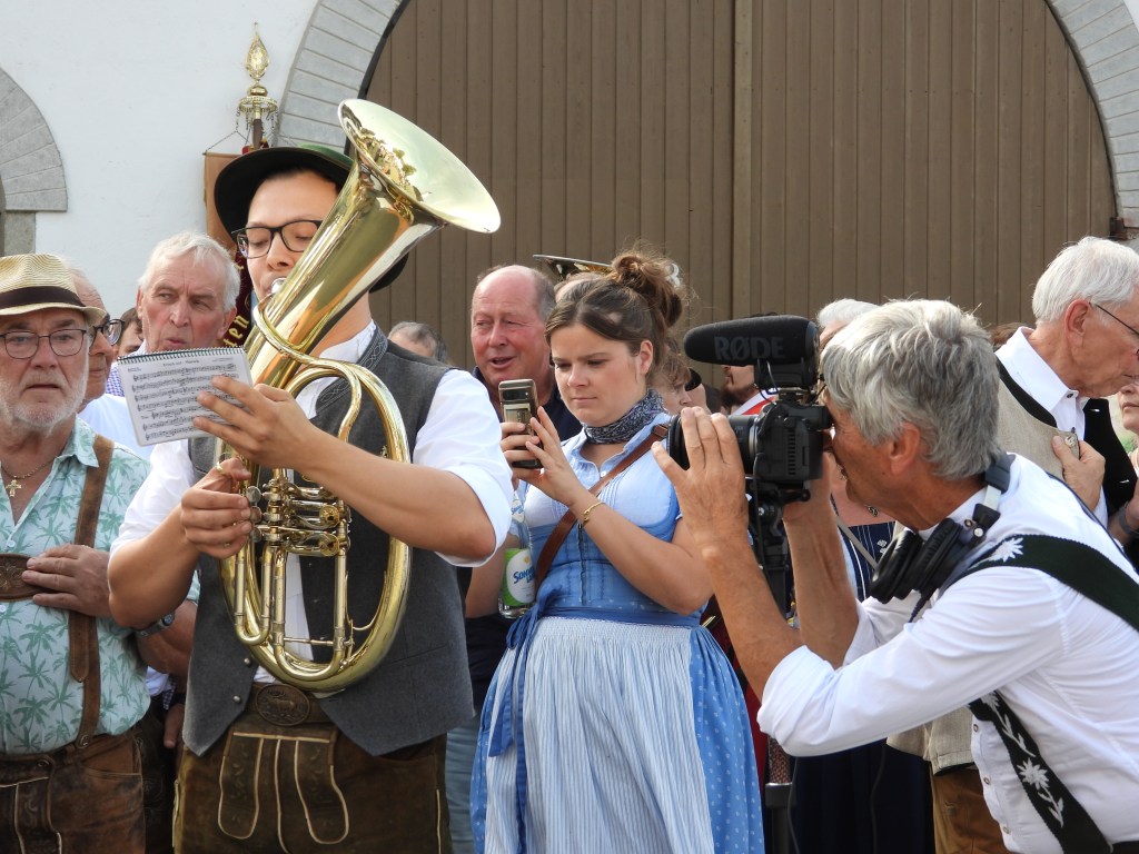 Eine Gruppe von Menschen bei einer Veranstaltung, während ein Musiker mit einer Tuba und Notenblättern spielt. Im Hintergrund sind weitere Teilnehmer in traditioneller Kleidung und eine Person, die ein Video aufnimmt.