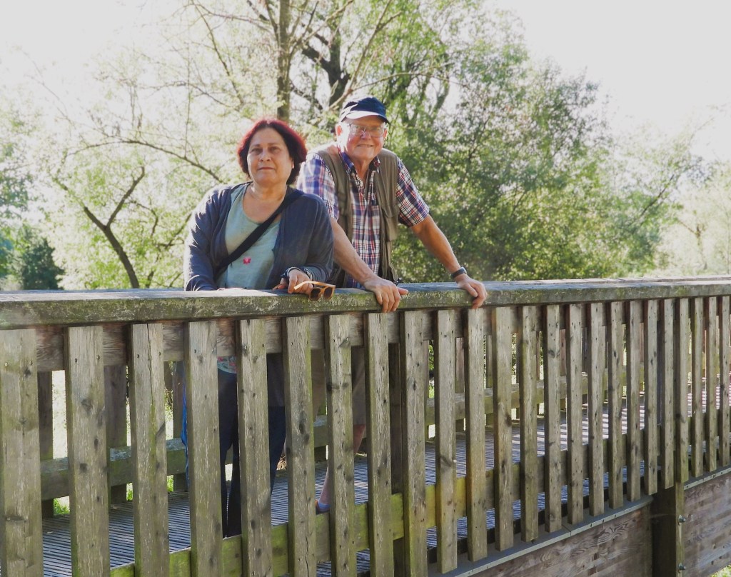 Ein Paar steht auf einer Holzbrücke, umgeben von Bäumen und natürlicher Landschaft, mit freundlichen Gesichtsausdrücken.