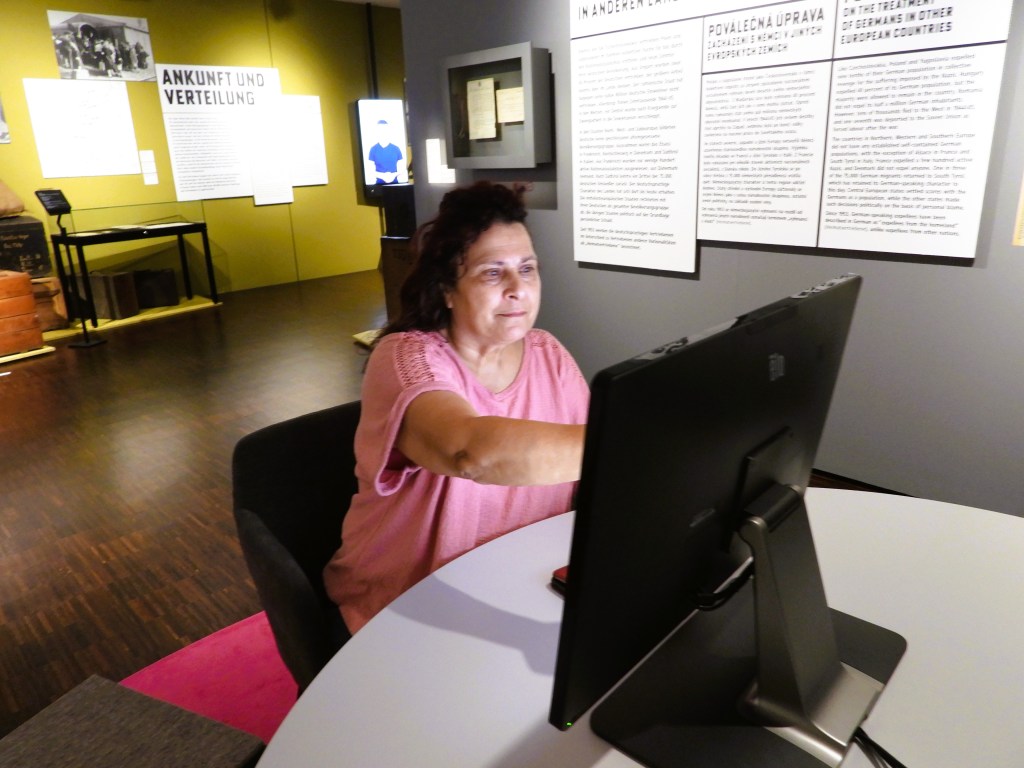 Eine ältere Frau sitzt an einem Tisch und arbeitet an einem Computer in einem Ausstellungsraum.