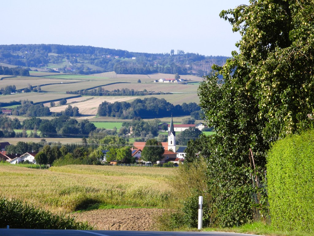 Blick auf eine ländliche Landschaft mit Feldern, Bäumen und einem kleinen Dorf, das eine Kirche mit einem spitzen Turm umfasst.