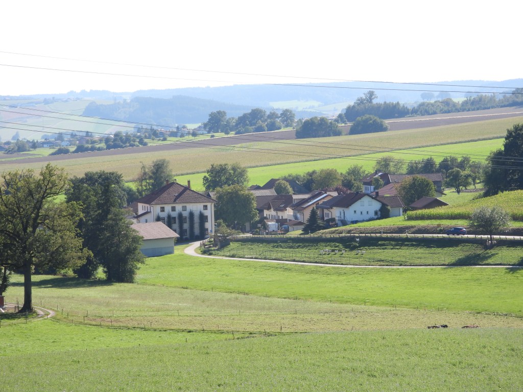 Landschaft mit einem kleinen Dorf, umgeben von Feldern und Hügeln, unter blauem Himmel.
