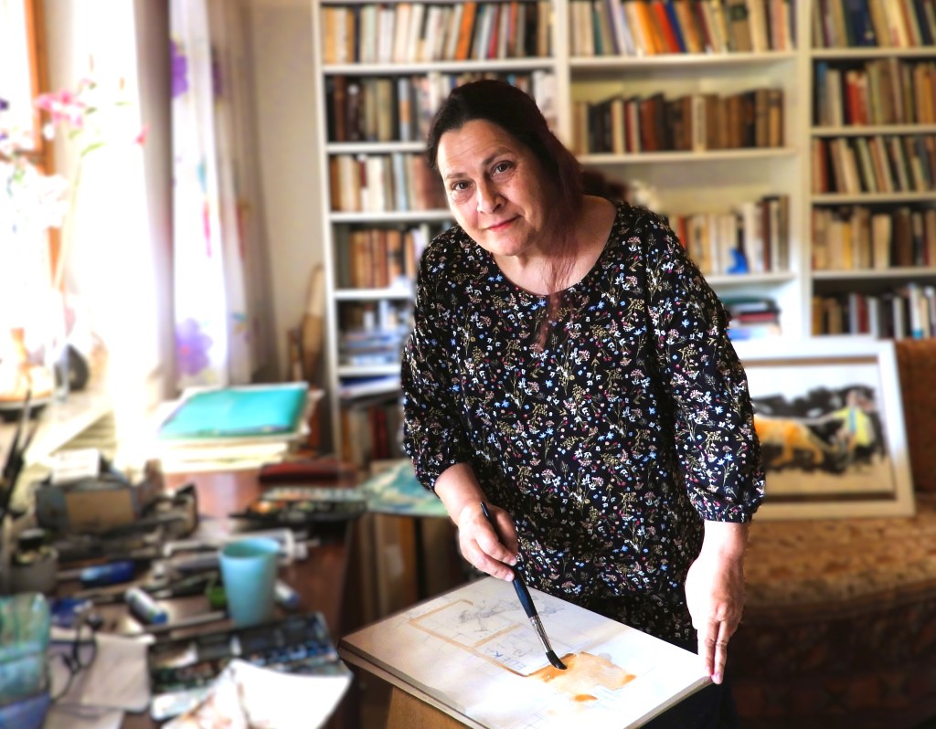 Eine Frau steht in ihrem Atelier und malt mit einem Pinsel auf einer Leinwand. Im Hintergrund sind Bücherregale und diverses Kunstmaterial zu sehen.