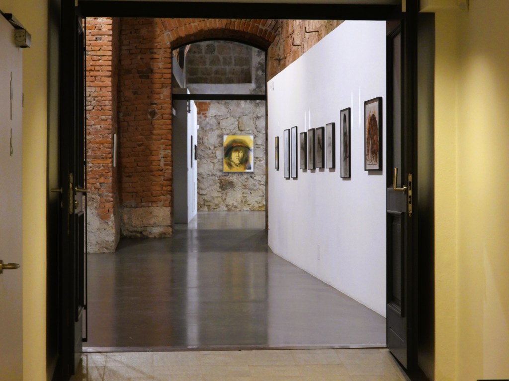 Blick auf einen Flur in einer Galerie mit einer Kombination aus Ziegel- und Steinwänden, in dem rahmende Kunstwerke an der Wand ausgestellt sind.