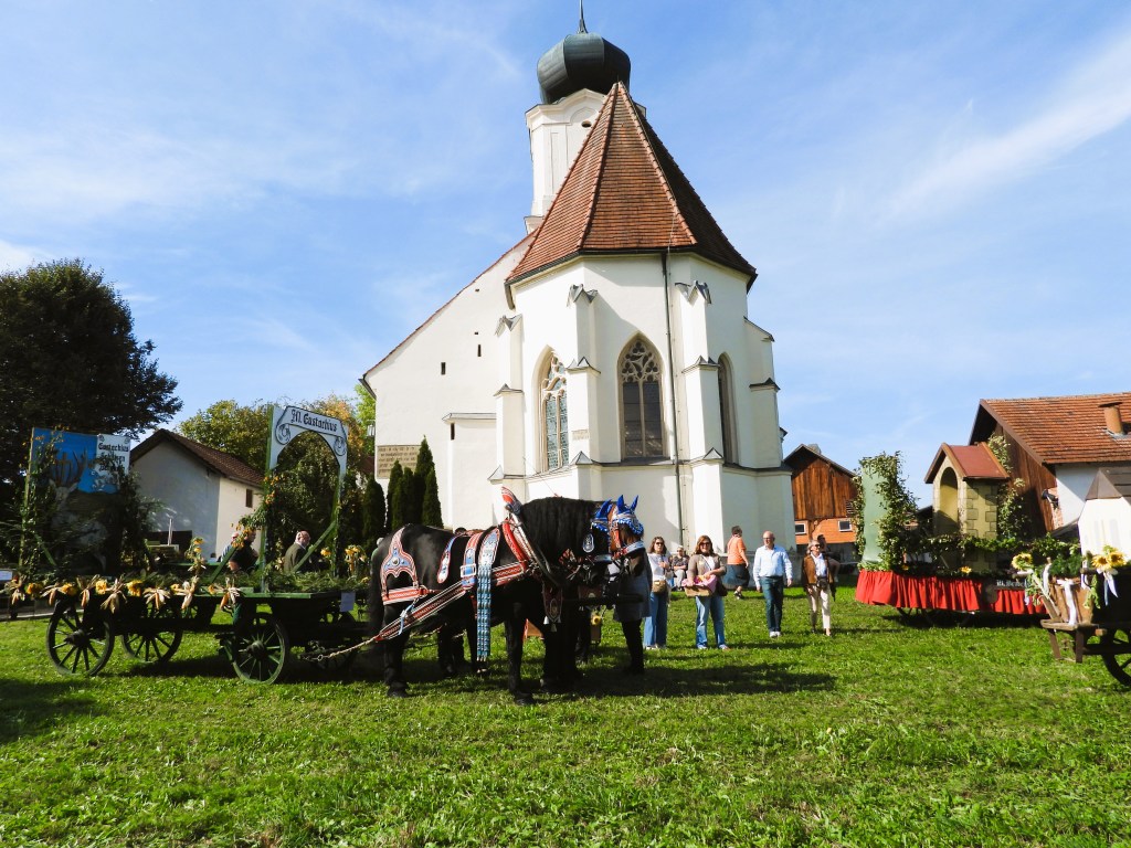 Eine Gruppe von Menschen steht vor einer Kirche, während ein Pferd mit einem festlich geschmückten Wagen auf einer Wiese steht.