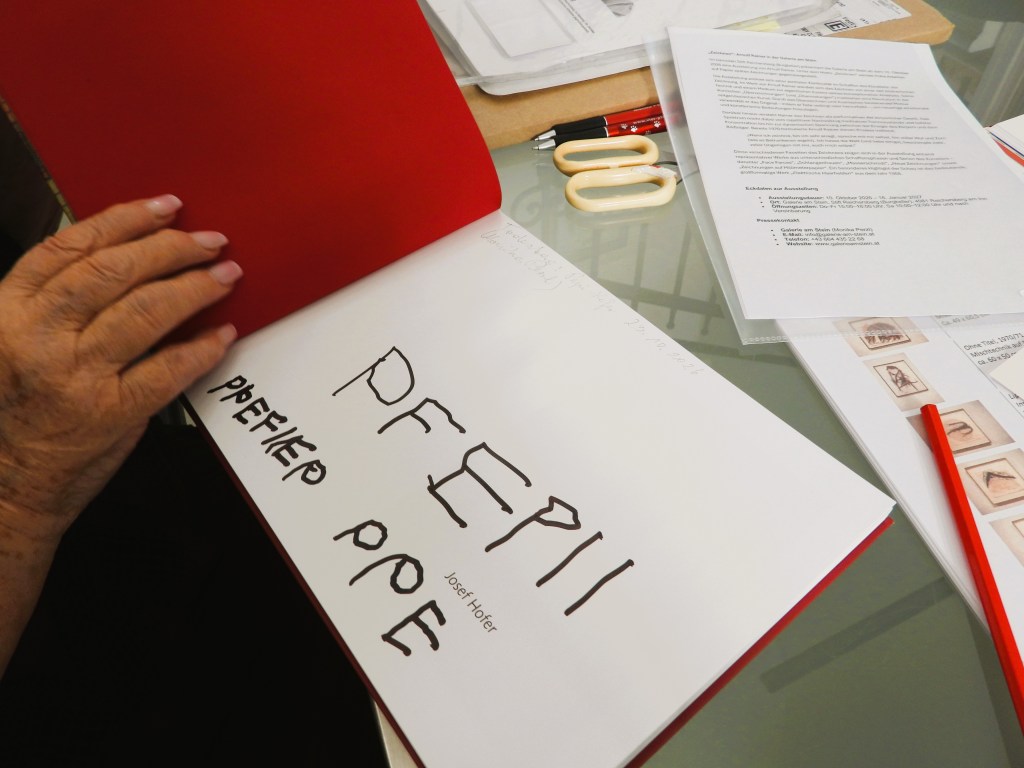 Eine Hand hält ein geöffnetes Buch mit handgeschriebenen Buchstaben 'PFEI', 'PFEHER' und 'Josef Hofer'. Um das Buch herum liegen Arbeitsunterlagen und Schreibutensilien auf einem Tisch.